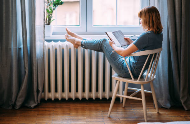 Jeune femme fait une pause lecture devant une fenêtre et à côté d'un chauffage