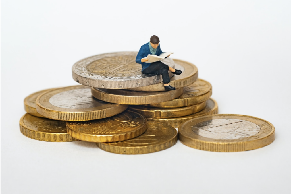 Une petite figurine en plastique est en train de lire sur un tas de pièces de monnaie, géantes par rapport à sa taille.