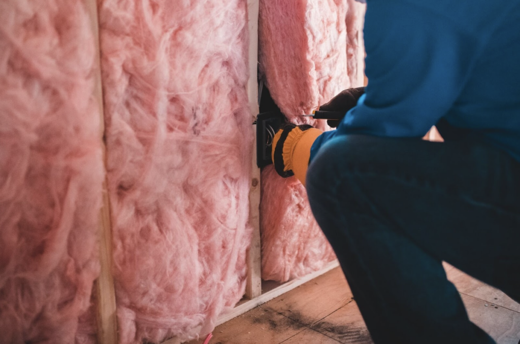 Un homme avec des gants noirs est en train d'installer de l'isolation en fibre de verre de couleur rose sur des murs.