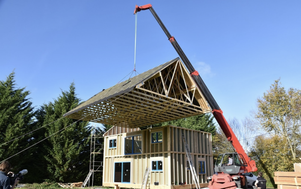 Maison en travaux avec un plan large, dans un milieu naturel semblable à une forêt. Une grue est en train de soulever un toit en élévation au-dessus d'une structure de maison en bois.