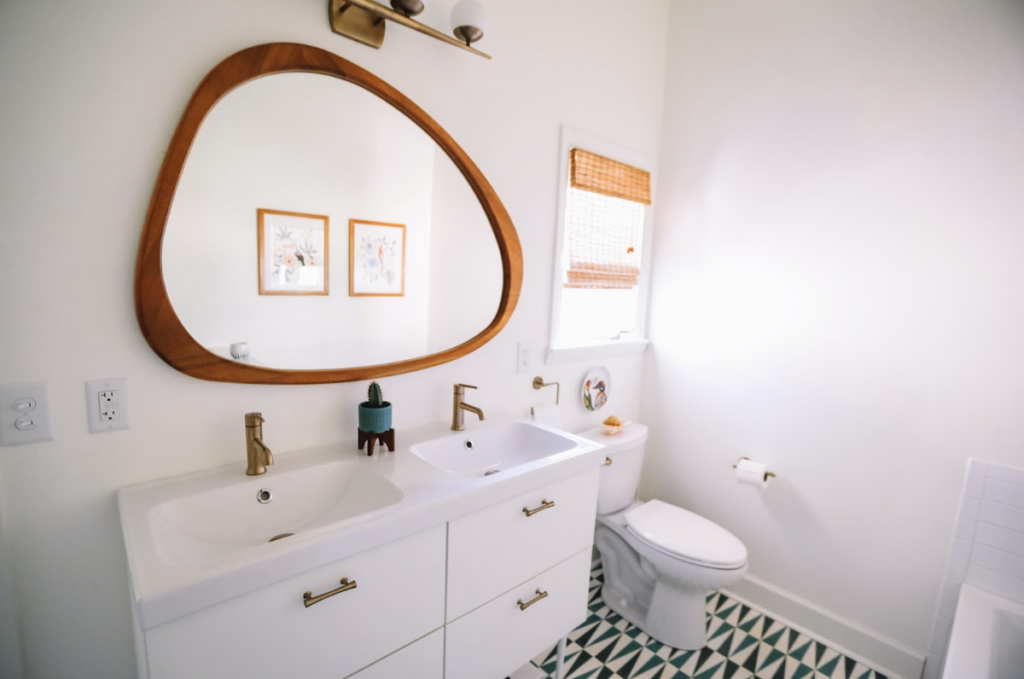Salle de bain neuve et moderne avec un grand miroir central. La pièce est claire et éclairée, avec des murs et des meubles blancs et une décoration très sobre. Seul le sol est coloré de motifs qui tranchent avec la sobriété de la pièce.