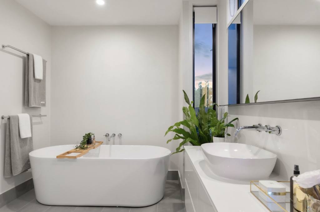 Grande salle de bain moderne et claire avec une baignoire et de grands lavabos blancs. C'est une salle de bain de luxe très éclairée et avec une grande plante dans le coin de la pièce.