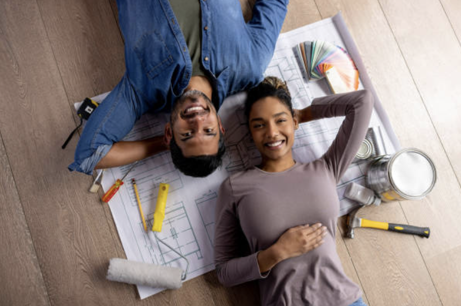 Un couple est allongé sur le dos sur son parquet par dessus des éléments de dessins et des papiers d'architecture. La photo est prise du dessus.