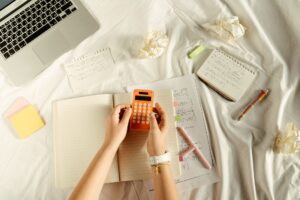 mains tenant une calculatrice orange sur un lit avec des cahiers et boules de papier en vrac
