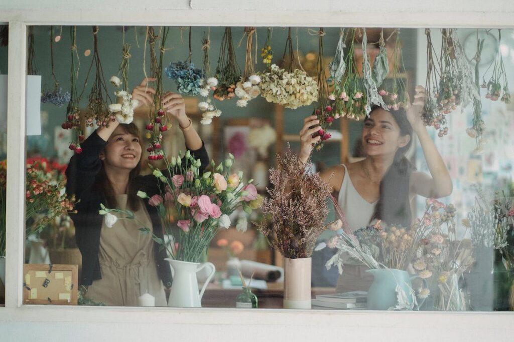 Une vitrine de magasin est en train d'être décorer par ses vendeuses. C'est très certainement un fleuriste car la vitrine n'est uniquement composé que de fleurs dans des vases ou alors suspendues.