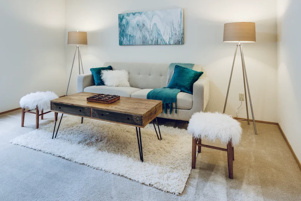 salon beige, deux lampes, un tapis beige avec une table basse en bois, un canapé blanc et des coussins bleu canard