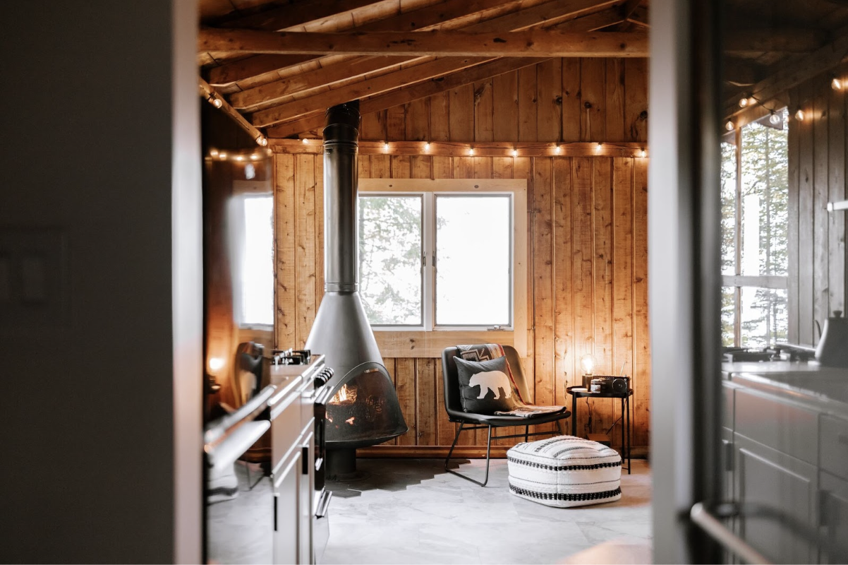 Intérieur de maison moderne avec des murs en bois, une cheminée en fonte, des guirlandes lumineuses, des coussins et de grandes fenêtres. C'est une pièce située dans des combles aménagés.