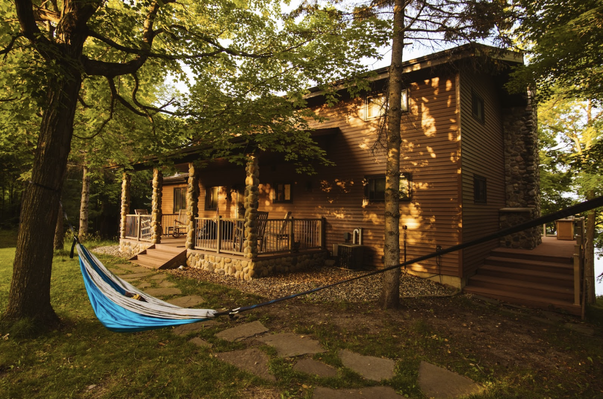 Maison en bois isolé en forme rectangulaire au milieu d'une forêt. Il y a un hamac à côté, l'image est assez sombre à cause de la couleur des arbres.